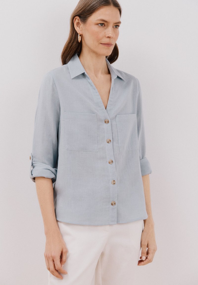 Camicia azzurro chiaro, con bottoni, realizzata in un tessuto morbido, con due tasche frontali, colletto e maniche arrotolate. Design classico e consistenza liscia.