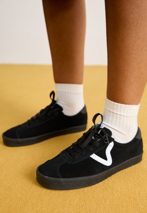 Zwarte suède sneakers met witte accenten, vetersluiting, ronde neus en een textuurrubberen zool, gedragen met geribbelde witte sokken op een gele achtergrond.