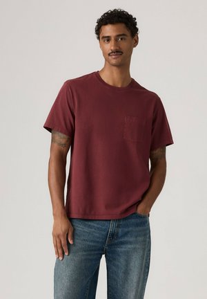 Uomo con capelli ricci e baffi che indossa una maglietta bordeaux a maniche corte e jeans blu, in piedi con una mano in tasca.