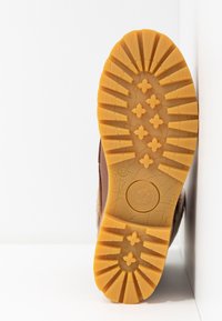 Semelle de bottine marron avec un motif en caoutchouc jaune texturé antidérapant, présentant des formes d'étoiles et de losanges, ainsi qu'un logo circulaire au centre.