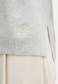 Maglione a maglia grigio chiaro con orlo e logo "LIU•JO MILANO" ricamato sopra il bordo a coste, abbinato a pantaloni in tessuto beige.