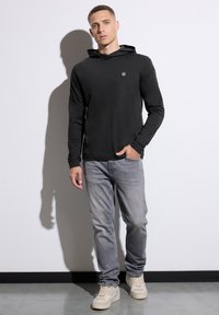 Black long-sleeve hoodie with a front logo detail, paired with light grey jeans and beige high-top sneakers on a smooth floor.