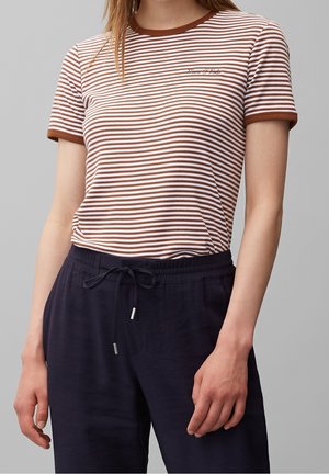 Kvinde iført brun- og hvidstribet kortærmet skjorte med brun kant og marineblå bukser med snor mod grå baggrund.