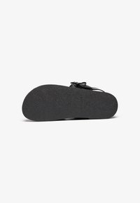 Svart sandal med en teksturert gummisåle, tynne stropper og dekorativ spenne. Sålen er flat og designet for komfort.
