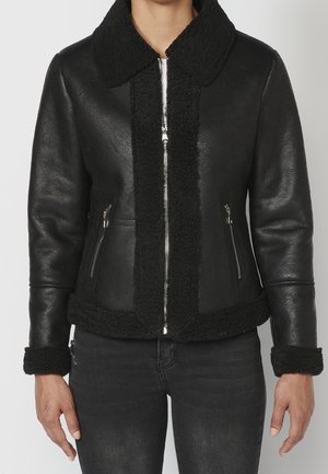 Czarna skórzana kurtka z kołnierzem, mankietami i dolnym wykończeniem z owczej wełny, z przodu z zamkiem błyskawicznym i zamykanymi kieszeniami bocznymi, noszona przez osobę w czarnych dżinsach.