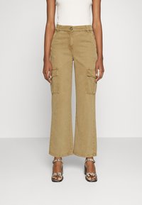 Pantalon cargo beige à jambes larges, avec deux poches frontales et une fermeture par bouton. Tissu en coton avec une texture douce. Associé à des talons à brides.