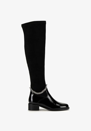Black over-the-knee boots featuring a suede upper and a glossy patent leather ankle. A silver chain accents the ankle area. Chunky heel design.