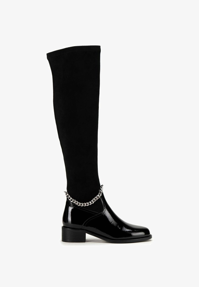 Botas negras por encima de la rodilla con parte superior de ante y tobillo de charol brillante. Una cadena plateada destaca en la zona del tobillo. Diseño de tacón grueso.