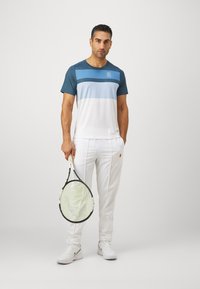 Mann i en fargeblokkert blå og hvit skjorte, hvite bukser og joggesko som står og holder en tennisracket med den ene hånden.