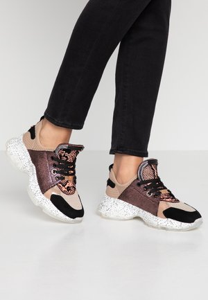 Jambes portant un pantalon noir et des baskets multicolores avec des semelles blanches tachetées, des accents en motif serpent or rose et noir, debout sur un sol clair.