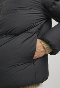 Schwarze Daunenjacke mit einer Seitentasche und gerippten beigen Bündchen, mit einer glatten Textur und leicht glänzender Oberfläche.