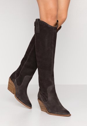 Bottes en daim brun foncé jusqu'au genou avec des talons en bois, portées sur des jambes croisées contre un fond clair.