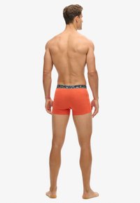 Bermudas naranjas para hombre, hechas de algodón, con una cinturilla elástica negra que presenta el nombre de la marca "SUPERDRY" repetido.