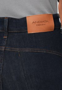 Tamni traperice s smešnim kožnim komadom na kojem su urezane riječi "ANDIATA" i "DENIM". Kontrastni šavovi i klasični dizajn s pet džepova.