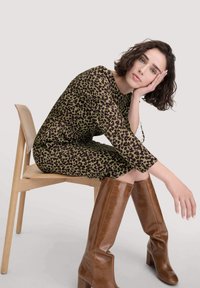 Langärmliges, figurbetontes Kleid in Dunkelgrün mit einem braunen Leopardenmuster. Knielange, glänzende braune Lederstiefel mit Blockabsatz. Modell sitzt auf einem Stuhl.