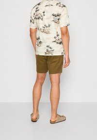 Korte mouwen, crèmekleurig shirt met palmboomprint, gecombineerd met olijfgroene shorts. Model draagt beige sandalen en kijkt van de camera weg.