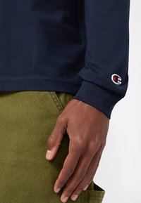 Dunkelhäutige Hand, die nahe der Tasche einer olivgrünen Hose ruht, trägt ein langärmliges, dunkelblaues Hemd mit einem kleinen Champion-Logo am Bund.