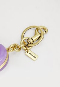 Violetti avaimenperä pyöreällä muotoilulla, kultaisilla yksityiskohdilla ja pienellä metallilapulla, jossa on kohokuvioitu "COACH". Suora pinta ja kiillotettu viimeistely.