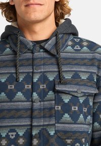 Navy fleece jacket with geometric patterns in teal and brown. Features a grey hood, snap buttons, and a chest pocket with black hardware.