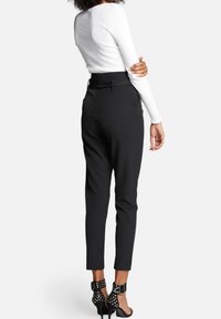 Mujer vistiendo pantalones negros ajustados, una blusa blanca de manga larga y sandalias de tacón alto negras con tachuelas, de pie con los brazos cruzados.