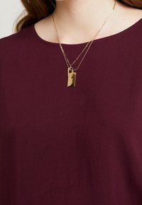 Burgunderrød top med rund halsudskæring, der har lagdelte guldkæder og en rektangulær vedhæng som detalje. Glat stofstruktur.