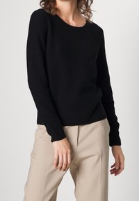 Person in einem schwarzen, langärmeligen, gerippten Pullover und beigen Hosen, der vor einem schlichten, hellen Hintergrund steht.