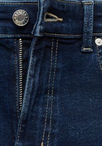 Jeans en denim bleu foncé en gros plan montrant une fermeture éclair métallique ouverte, un bouton métallique avec texte gravé, et une passants de ceinture avec une couture beige.