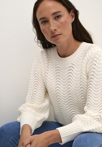 Pull en tricot blanc avec un motif en vagues, présentant des poignets côtelés et un col rond. Le mannequin est assis avec un jean en denim clair.