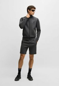 Mörkgrå träningsjacka och matchande shorts i slät tyg. Detaljer inkluderar dragkedjestängning och logotyp på bröstet. Svarta sneakers och strumpor.
