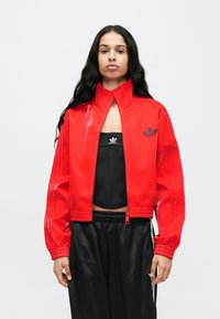 Rdeča vinilna krajšana jakna z visokim ovratnikom, z rebrastimi manšetami in majhnim črnim logotipom Adidas na levi strani prsnega koša.