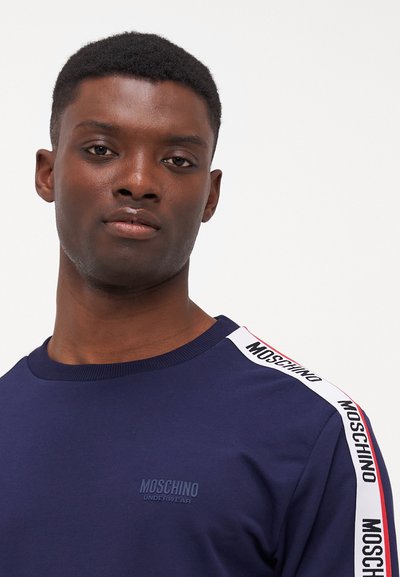 Tamsiai mėlynas ilgomis rankovėmis marškinis su apvaliu iškirpimu, tekstūra ir Moschino logotipo detalėmis baltose ir raudonose šoninėse juostose.