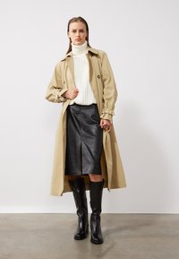 Cappotto trench beige con maniche lunghe, design doppiopetto. Maglione bianco a collo alto a costine, gonna a matita nera in pelle e stivali neri fino al ginocchio.