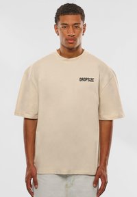 Beige oversized t-shirt lavet af bomuld, med korte ærmer og rund halsudskæring, og med et sort "DROPSIZE" logo på venstre bryst.
