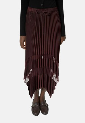 Kobieta w burgundowej plisowanej spódnicy midi z asymetrycznym dołem i czarnych mokasynach, w połączeniu z czarną bluzką z długim rękawem.