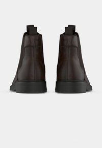 Un paio di stivaletti alla caviglia in pelle marrone scuro con lati elastici neri e linguette nere, mostrati da dietro su uno sfondo bianco.