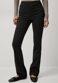 Zwarte, elastische broek met een gladde textuur en uitlopende benen, met een elastische tailleband. Draagt met zwarte platte schoenen.