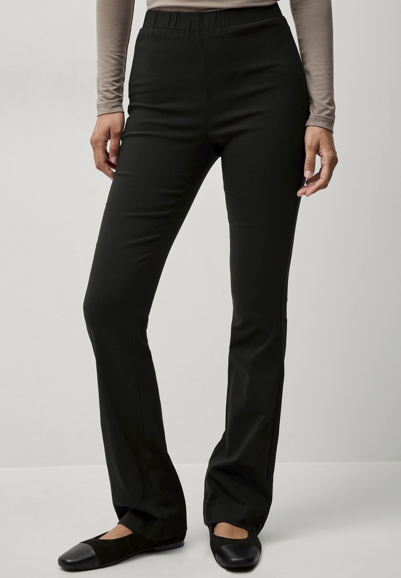 Zwarte, elastische broek met een gladde textuur en uitlopende benen, met een elastische tailleband. Draagt met zwarte platte schoenen.