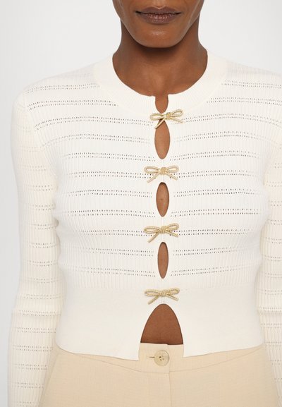 Hvit strikket cardigan med rund hals, med gull sløyfe detaljer og utskårne detaljer. Myk tekstur og ribbet design.