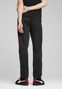 Schwarze Sporthosen mit einer figurbetonten Passform verfügen über bordeauxfarbene Seitenstreifen und elastische Bündchen. Das Modell trägt schwarze und rote Schuhe mit Klettverschluss.