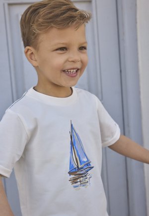 Camiseta blanca de manga corta con una ilustración de un velero azul en el frente, mostrada con un niño sonriente sobre un fondo azul.