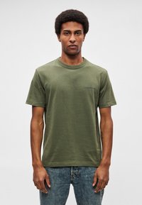 Oliivinvihreä puuvillainen t-paita pyöreällä pääntiellä ja lyhyillä hihoilla, jossa pieni tummansininen logo rinnassa. Karkea kangas ja rento istuvuus.