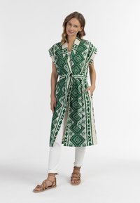 Kimono lungo verde e crema con un motivo geometrico, maniche corte, cintura con fiocco e tasche laterali, indossato sopra pantaloni bianchi e sandali.