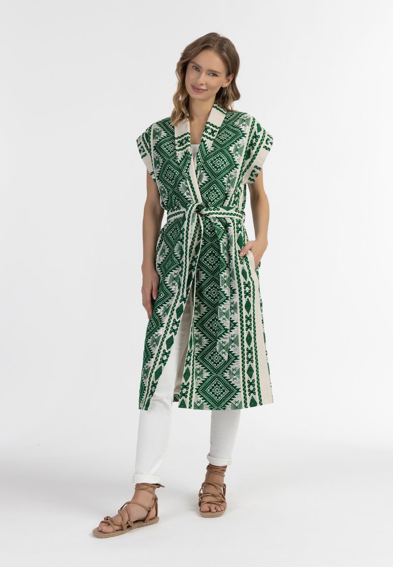 Kimono lungo verde e crema con un motivo geometrico, maniche corte, cintura con fiocco e tasche laterali, indossato sopra pantaloni bianchi e sandali.