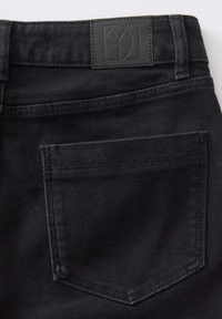 Jean en denim noir montrant la poche arrière et la ceinture avec un patch en cuir noir cousu.
