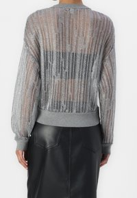 Srebrny, metaliczny sweter z siateczki z długimi, bufkowatymi rękawami, szarymi ściągaczami na mankietach i dole oraz przezroczystym wzorem, przez który widać czarny element garderoby pod spodem.