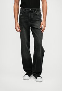 Jeans de ganga pretos, corte folgado, cinco bolsos, fecho de botão, textura de lavagem subtil, combinados com ténis pretos e uma camisa preta simples.