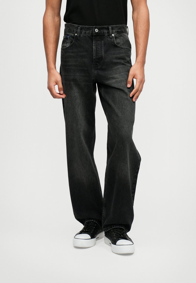 Jeans de ganga pretos, corte folgado, cinco bolsos, fecho de botão, textura de lavagem subtil, combinados com ténis pretos e uma camisa preta simples.