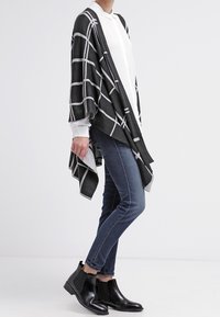 Envuelta de cuadros en blanco y negro con mangas sueltas, sobre una camisa blanca. Combinada con jeans ajustados y botas de tobillo negras.