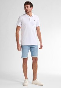 Polo bianco con un piccolo logo, pantaloni corti in denim azzurro arrotolati, e sneakers color crema. Outfit estivo semplice e casual.