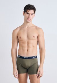Boxeri de culoare verde măslin cu un brâu albastru marin pe care este inscripționat brandul "BOSS" cu litere albe. Material neted și design ajustat.